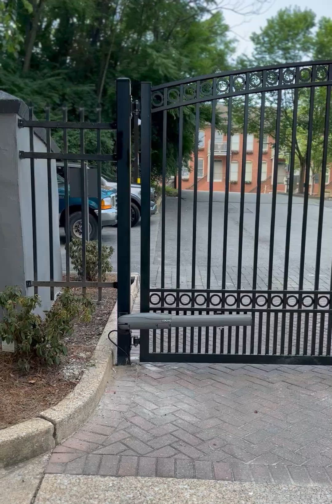 Black metal gate across a brick driveway, with trees, a parked car, and an orange building beyond.