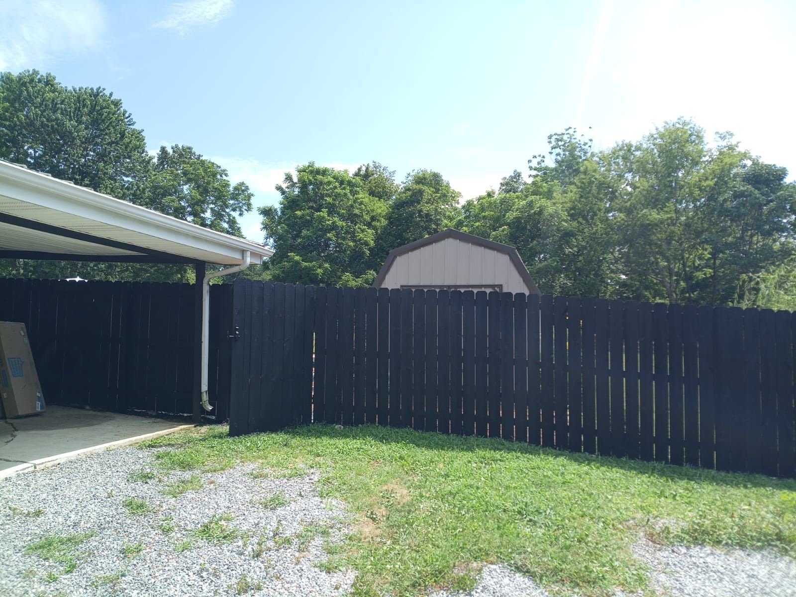 Backyard with black fence, gravel and grass, carport on the left, and a house roof behind trees.