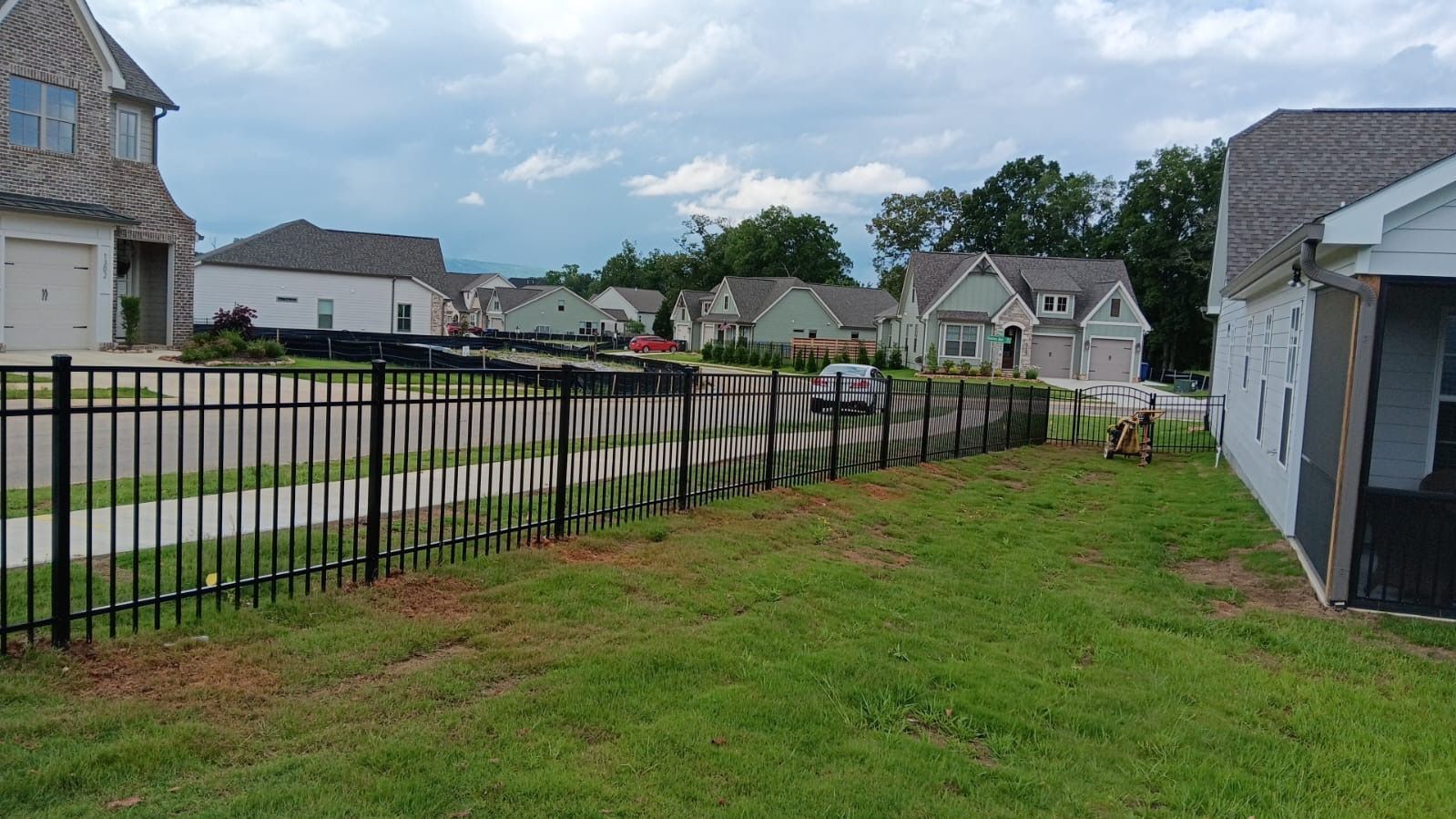 Suburban neighborhood with houses, black metal fence, green lawn, and cloudy sky
