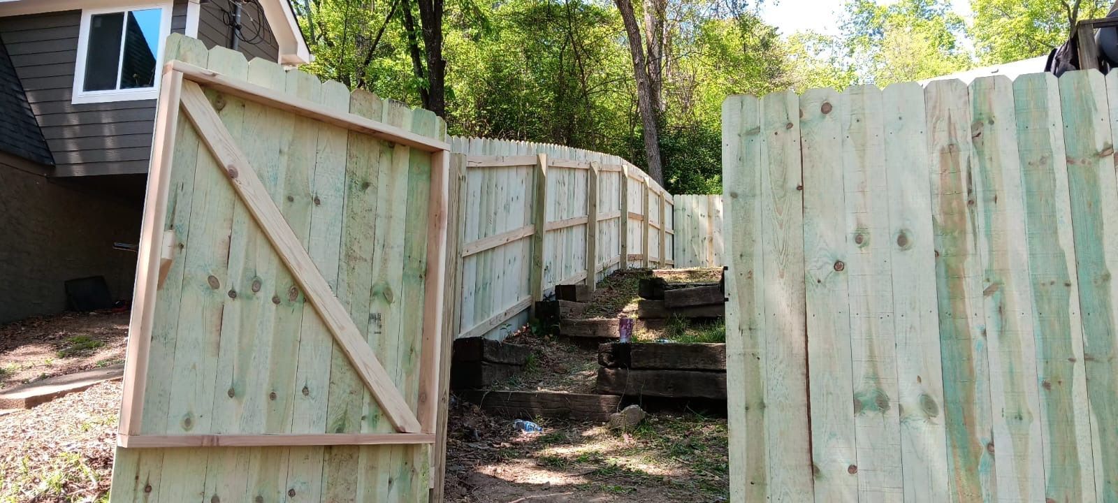 Wooden fence panels lining a narrow dirt path beside a house in a wooded yard