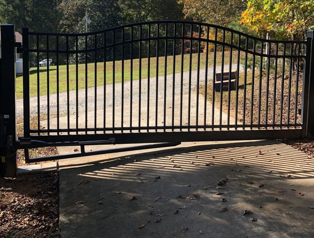 Black metal driveway gate across a sunlit concrete path with trees and fallen leaves behind it