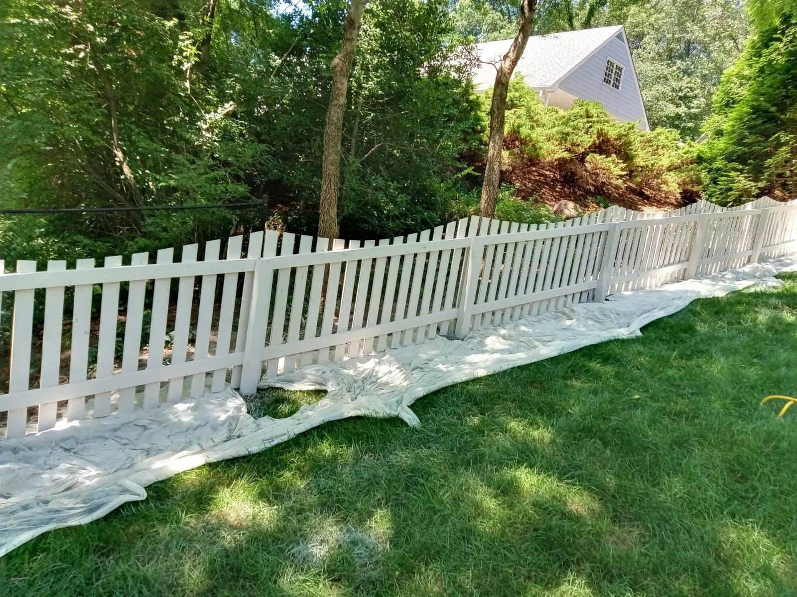White picket fence lining a green yard with white landscape fabric and trees in the background