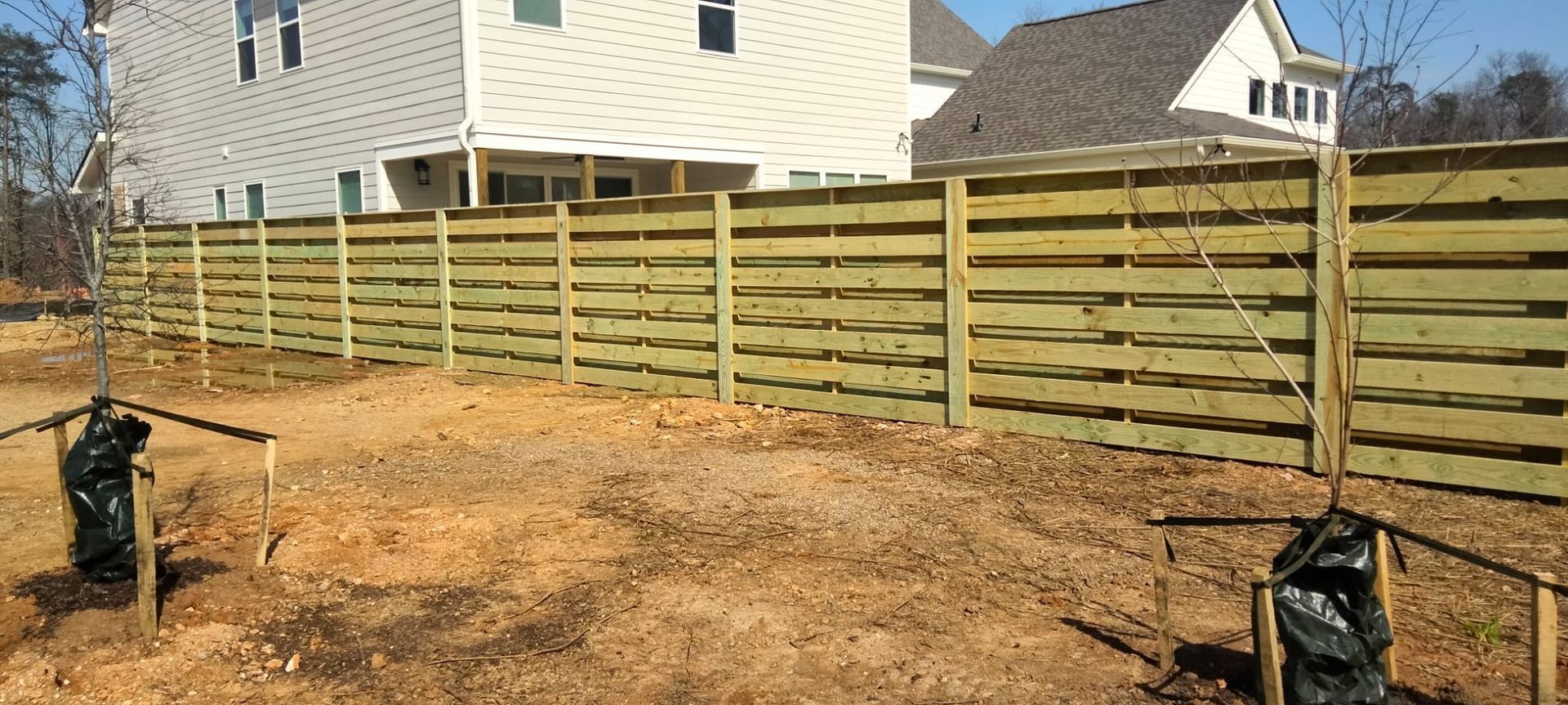 Backyard with a long wooden privacy fence and dirt yard beside a house, with small trees under construction bags