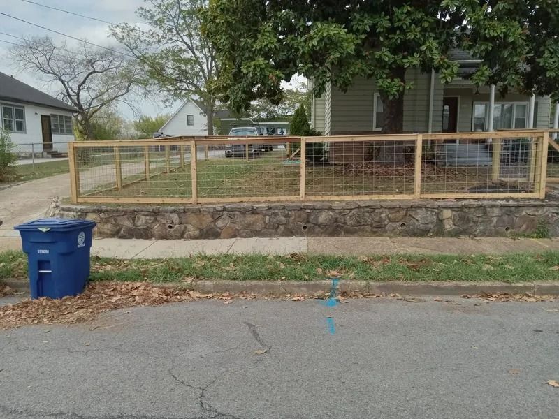 Residential sidewalk with blue recycling bin, stone retaining wall, and fenced front yard.