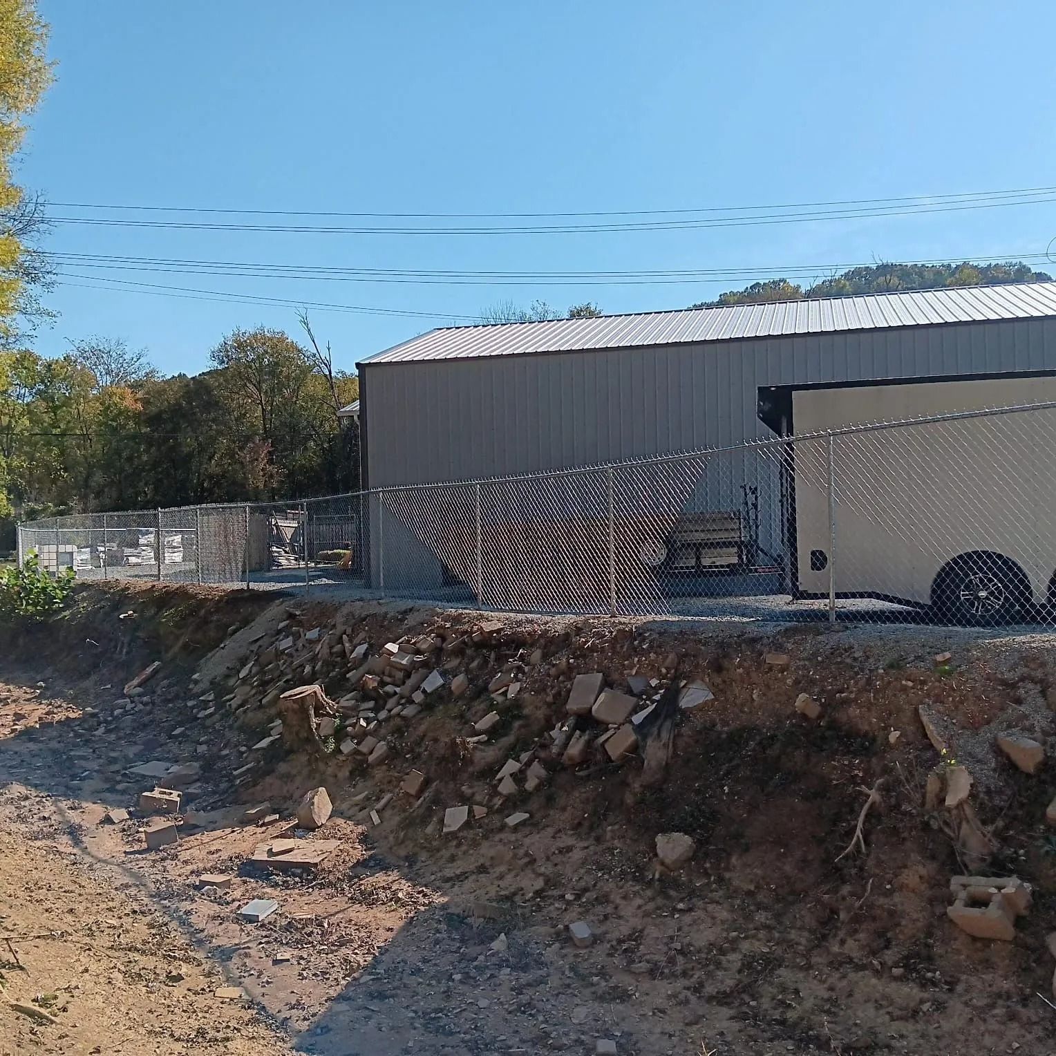 Rural dirt road beside a gray metal warehouse, with a chain-link fence and rocky embankment under a clear blue sky