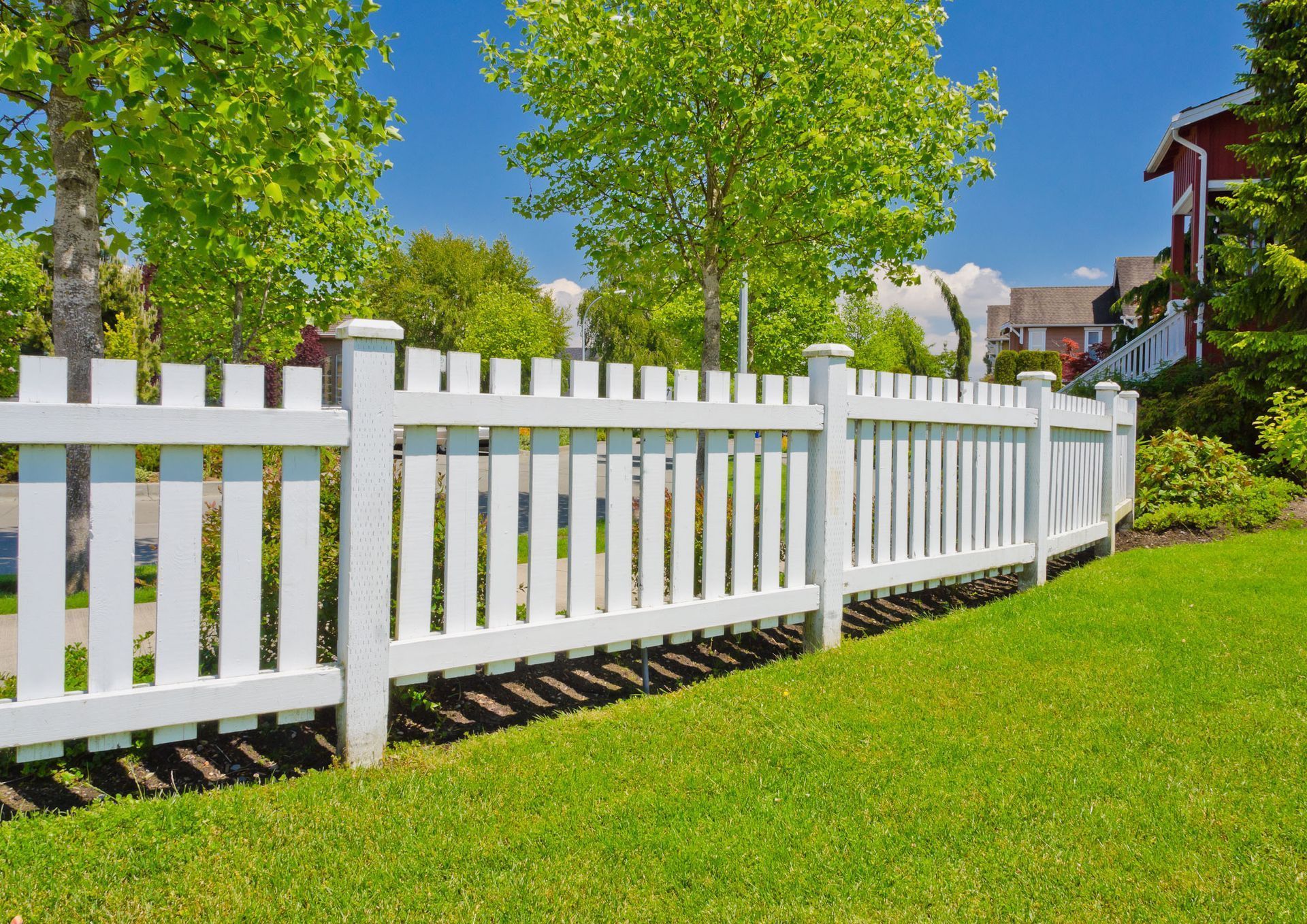 White picket fence along a sunny green lawn with trees and a house in the background