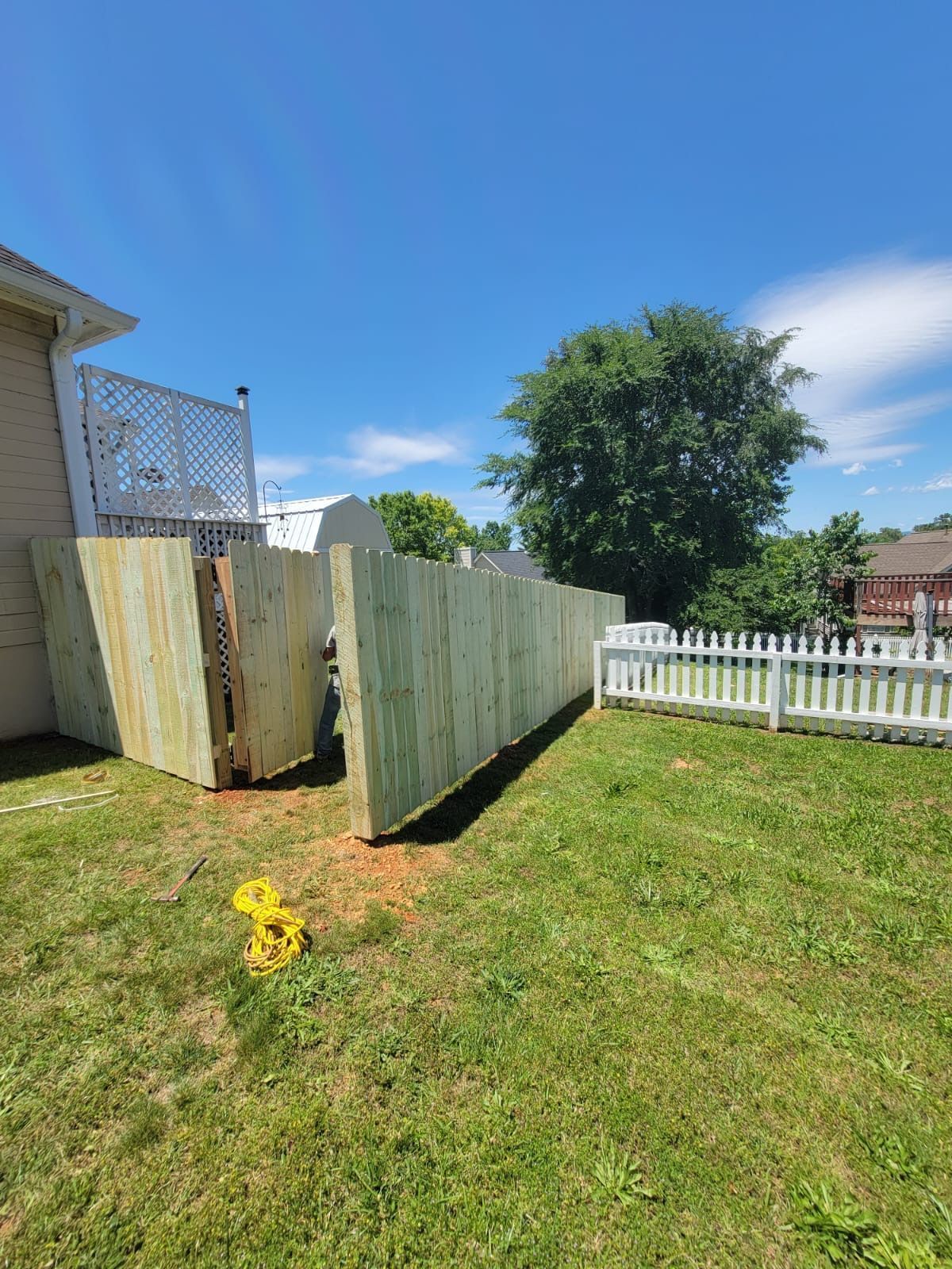 Backyard with a leaning wooden fence, green lawn, white picket fence, and a tree under a blue sky