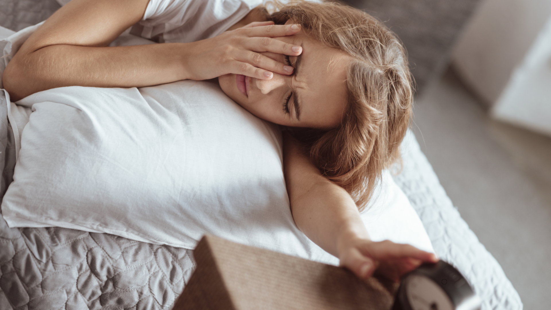 A woman is laying in bed covering her face with her hand while holding an alarm clock.