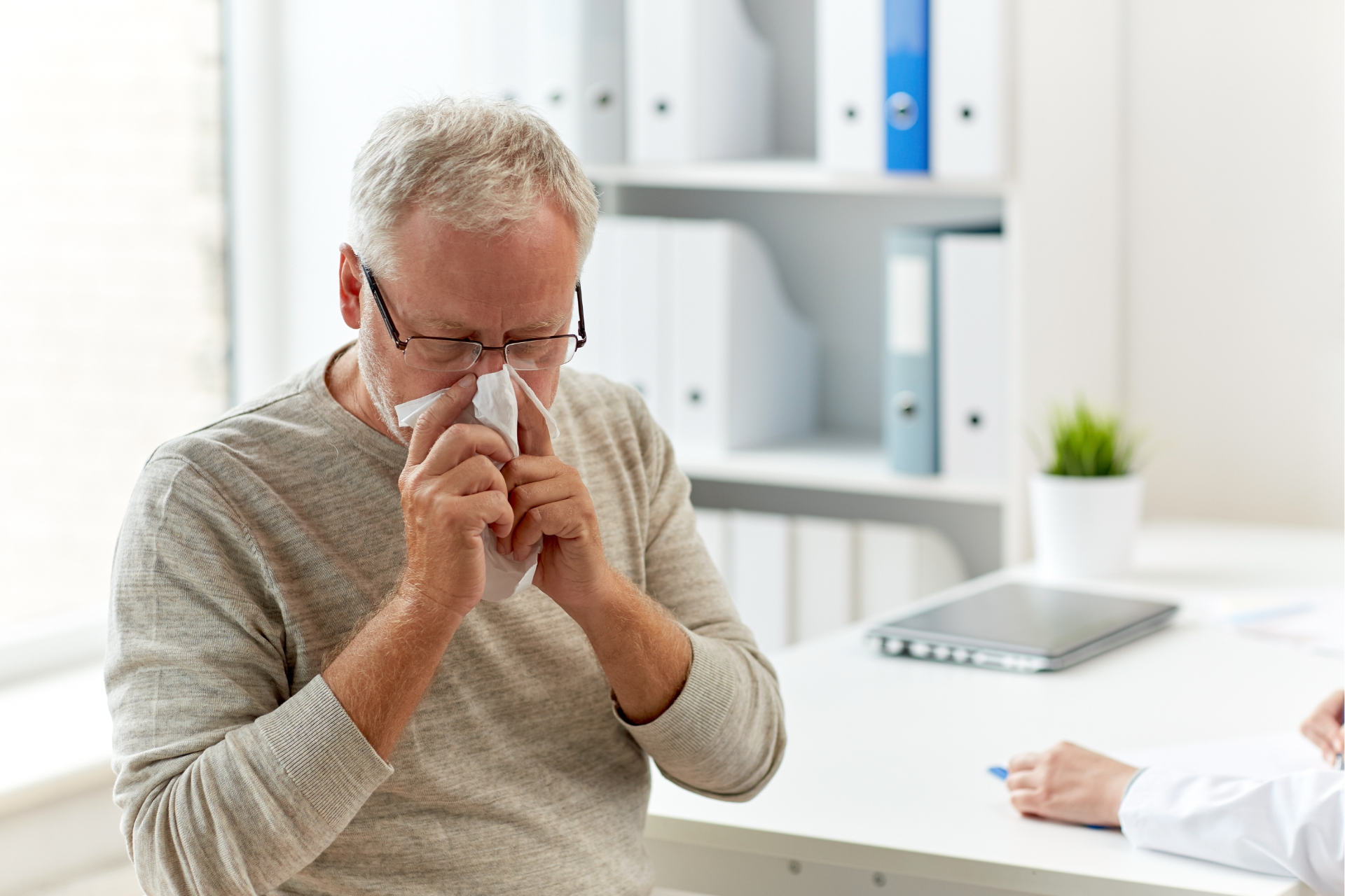 An elderly man is blowing his nose into a napkin while talking to a doctor.