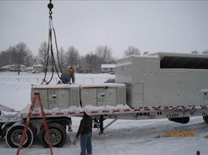 Workers loading equipment onto a trailer in a snowy field. A crane and ladder are present.