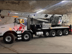 Crane truck under an archway; white cab with American flag art, extended boom.