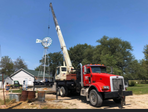 A crane lifts a windmill in a rural setting. A red truck, blue sky, and a house are visible.