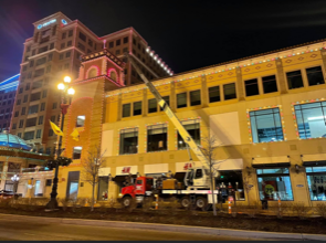 A crane in front of a brick building at night. A worker in an orange vest is visible.