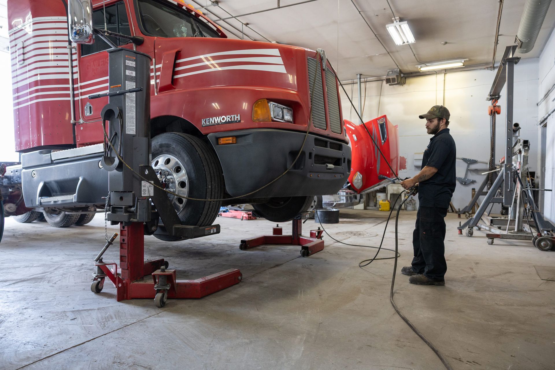 A mechanic works on a red semi-truck in a garage. The truck is raised on jacks, and a detached part lies nearby.
