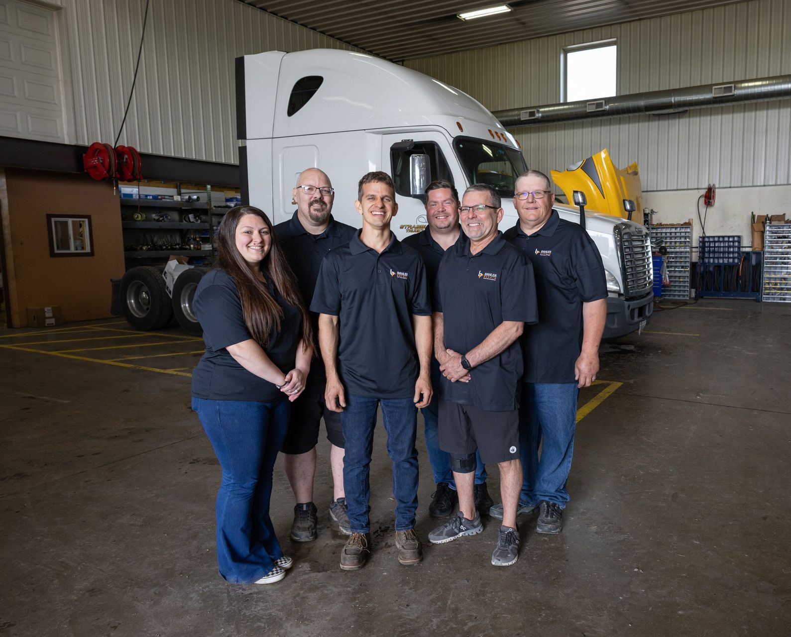 A group of seven people posing in front of a white semi-truck inside a garage. They are smiling, and appear to be a work team.