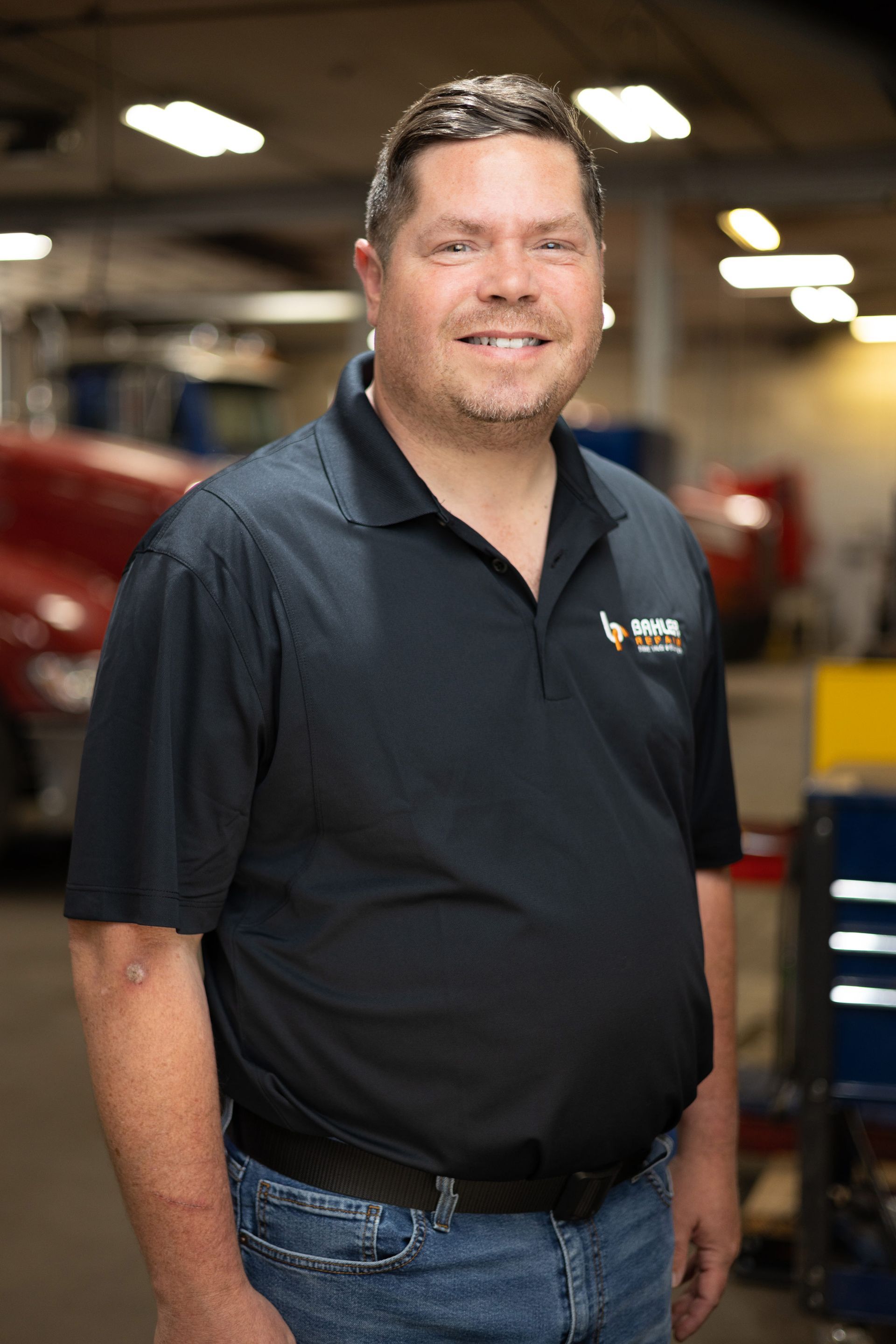 Man in a black polo shirt and jeans smiles in a garage. He has short brown hair and a logo on his shirt.