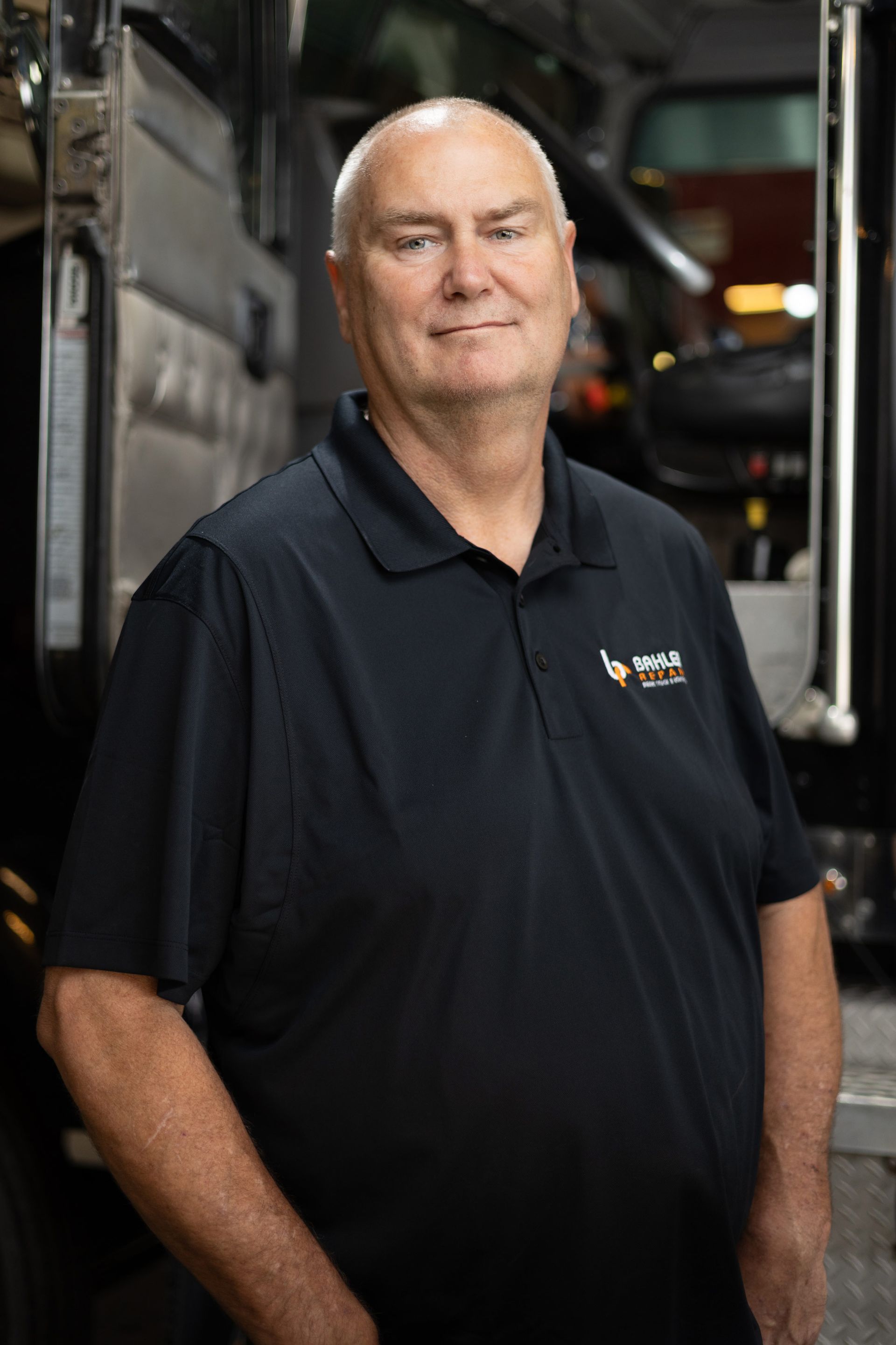 A middle-aged man in a black polo shirt, smiling, standing near a truck. He is in what appears to be a garage setting.