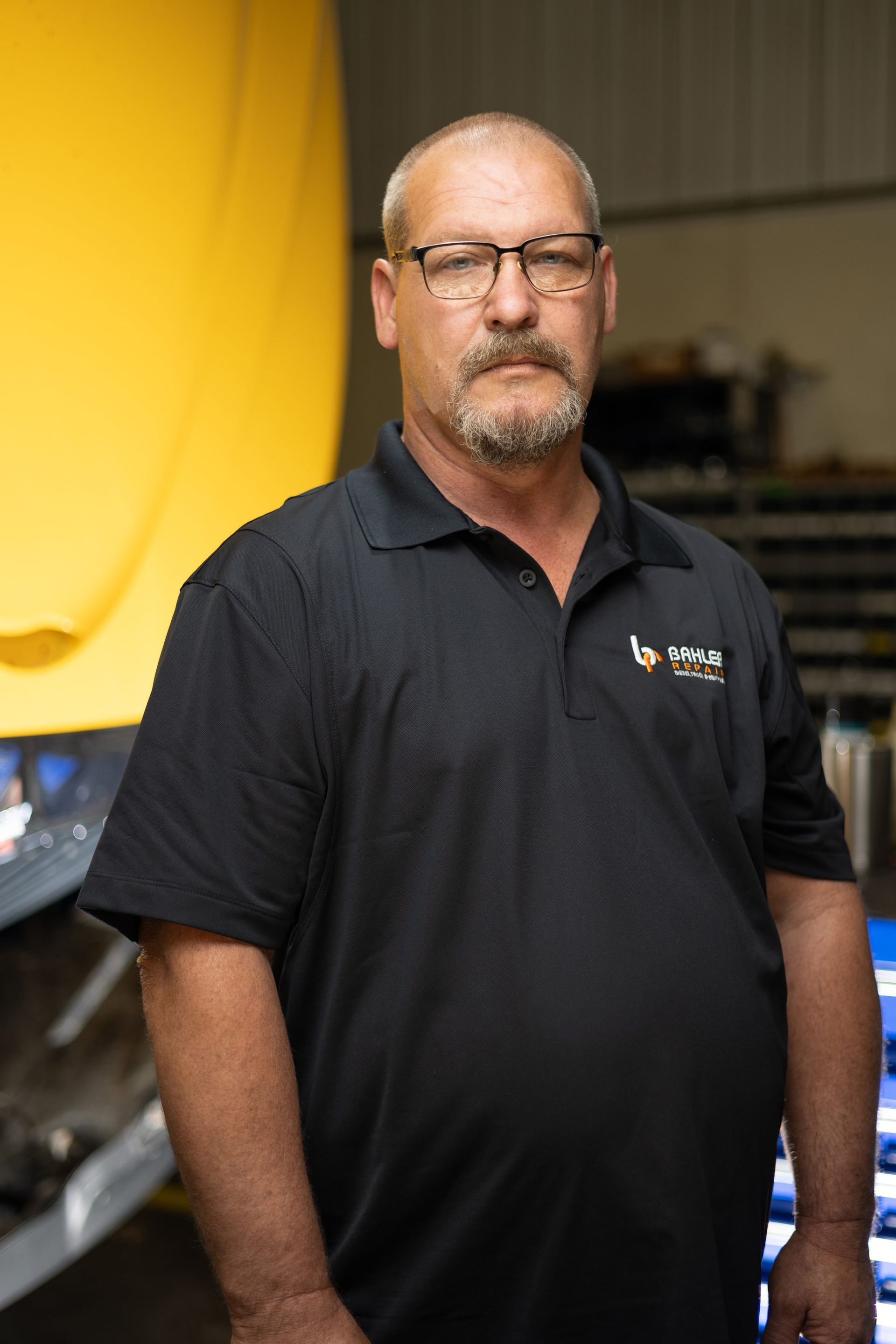 Man in a black polo shirt and glasses stands in a shop with yellow machinery, looking at the viewer.