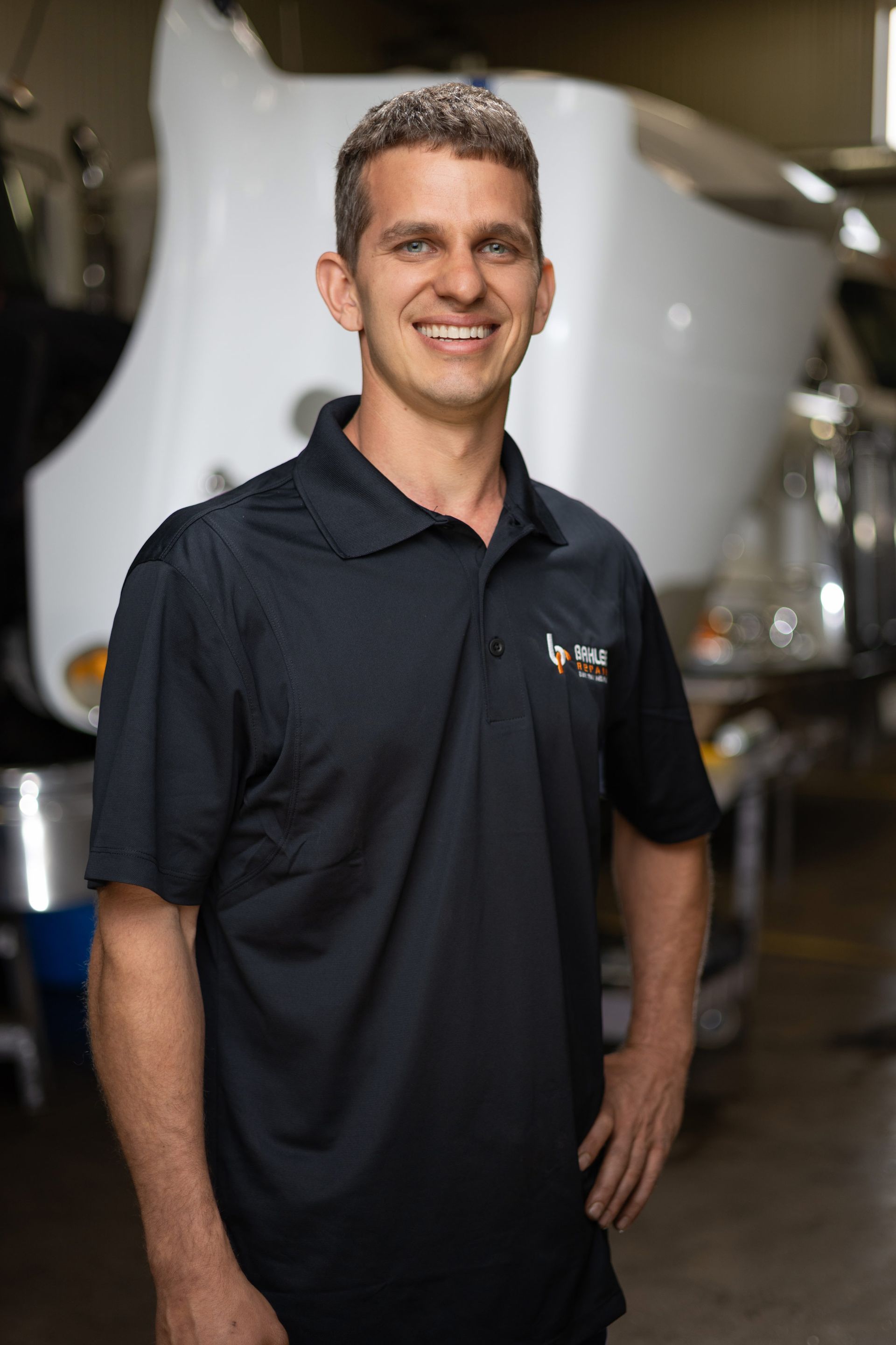 Smiling man in black polo shirt poses in front of a partially visible white vehicle; he's in a repair shop.