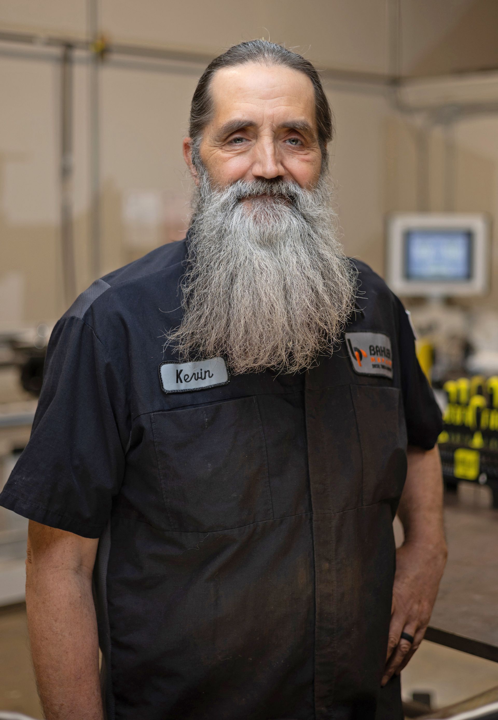 Man with long beard in work uniform, standing inside a workshop; smiling.