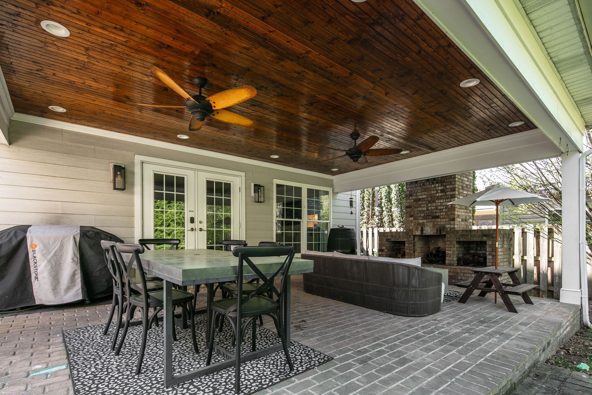 Covered patio with a dining set, gray stone floor, dark wood-paneled ceiling, ceiling fans, and an outdoor fireplace.