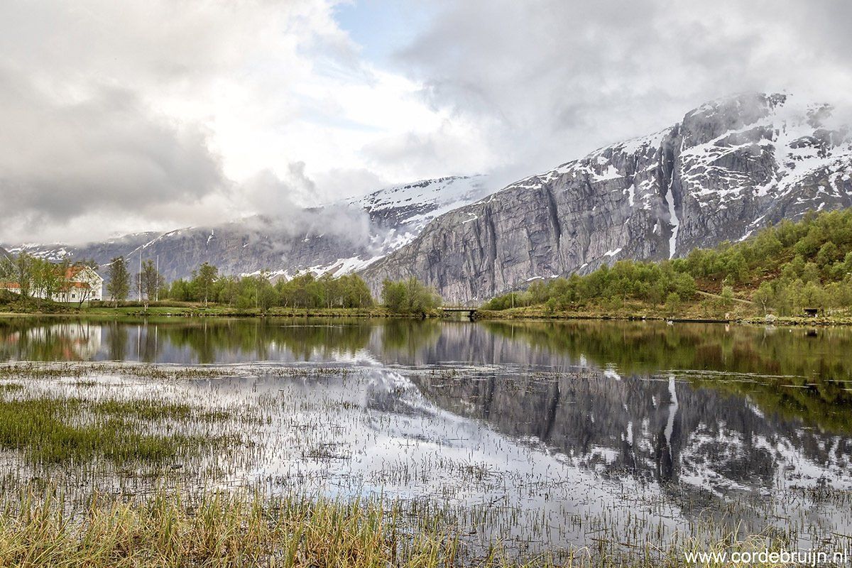 Lake reflects a snow-capped mountain under a cloudy sky. Trees line the shore.