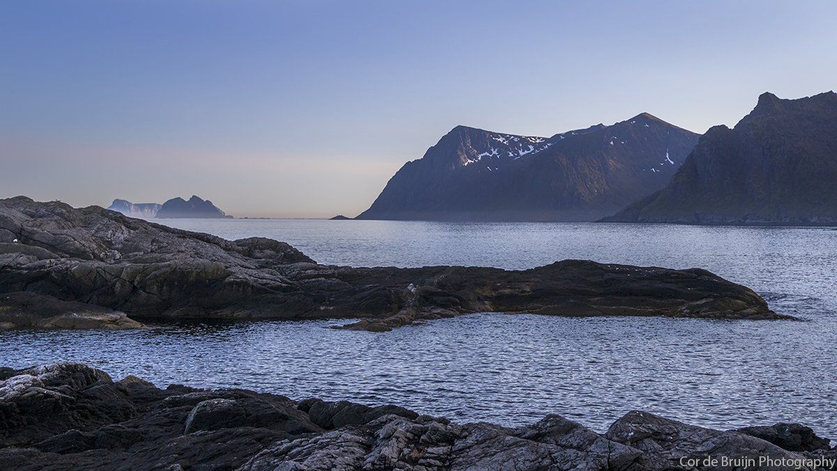 Rocky coastline with a calm sea reflecting a mountainous backdrop under a pale blue sky.