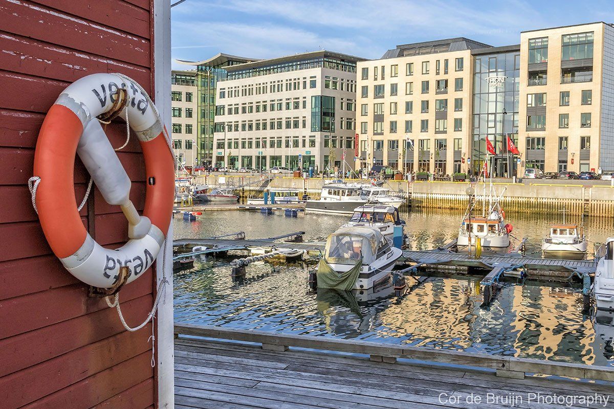 Orange and white life ring hangs on red wall, overlooking boats in a harbor, with beige buildings in the background.