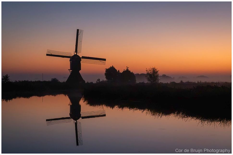 Windmill silhouetted at sunrise, reflected in calm water under an orange sky.