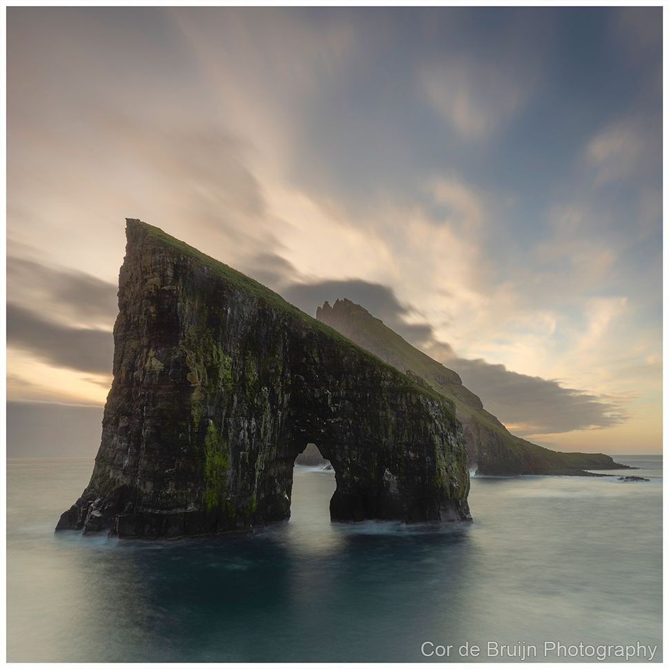 Rocky sea arch covered in greenery at sunset, with calm water and cloudy sky.