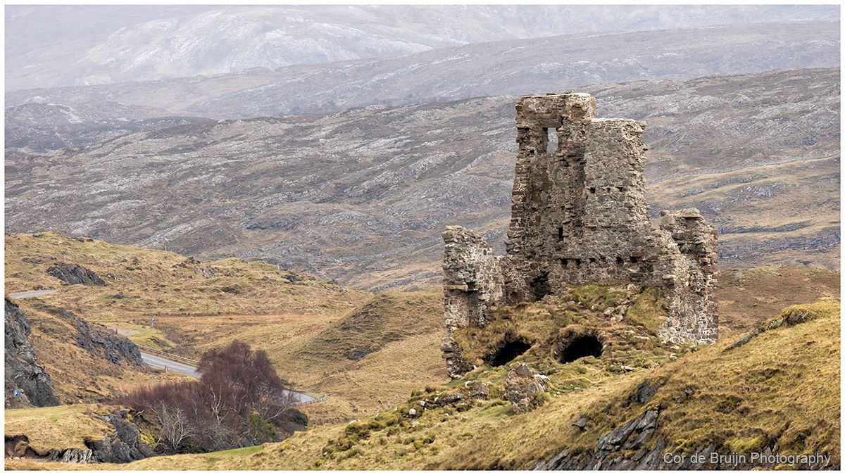 Ruined stone tower on a grassy hillside overlooking a wide valley under a cloudy sky