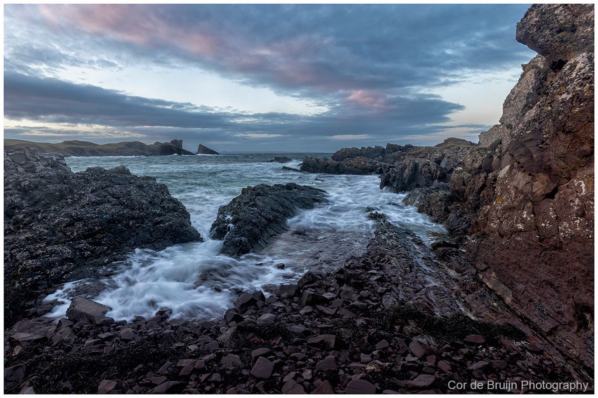 Rocky seaside cove at dusk with waves washing between dark rocks under a cloudy sky