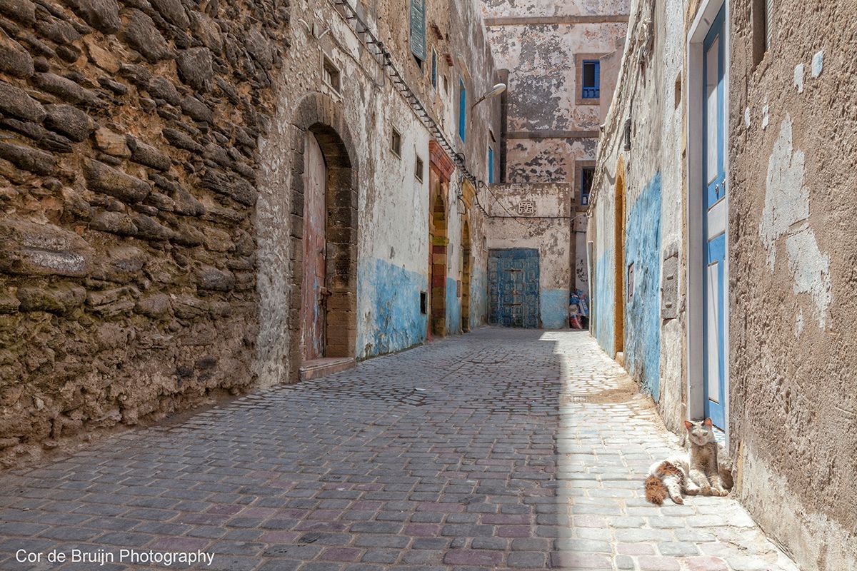 Narrow cobblestone street in a Moroccan city, lined with weathered stone and stucco buildings.