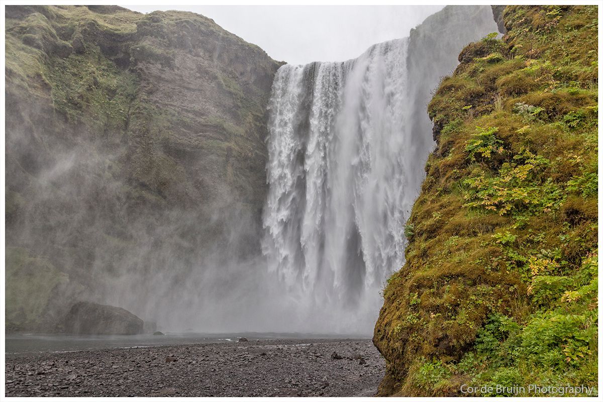 Waterfall cascading down a cliff face, creating mist. Rocky foreground, green vegetation on the right.
