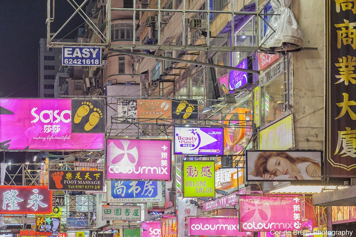Night view of dense, illuminated shop signs in Hong Kong. Pink, purple, yellow lights; text in Chinese and English.