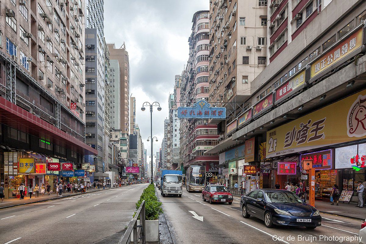 Street scene in Hong Kong with tall buildings, shops, traffic, and cloudy sky.