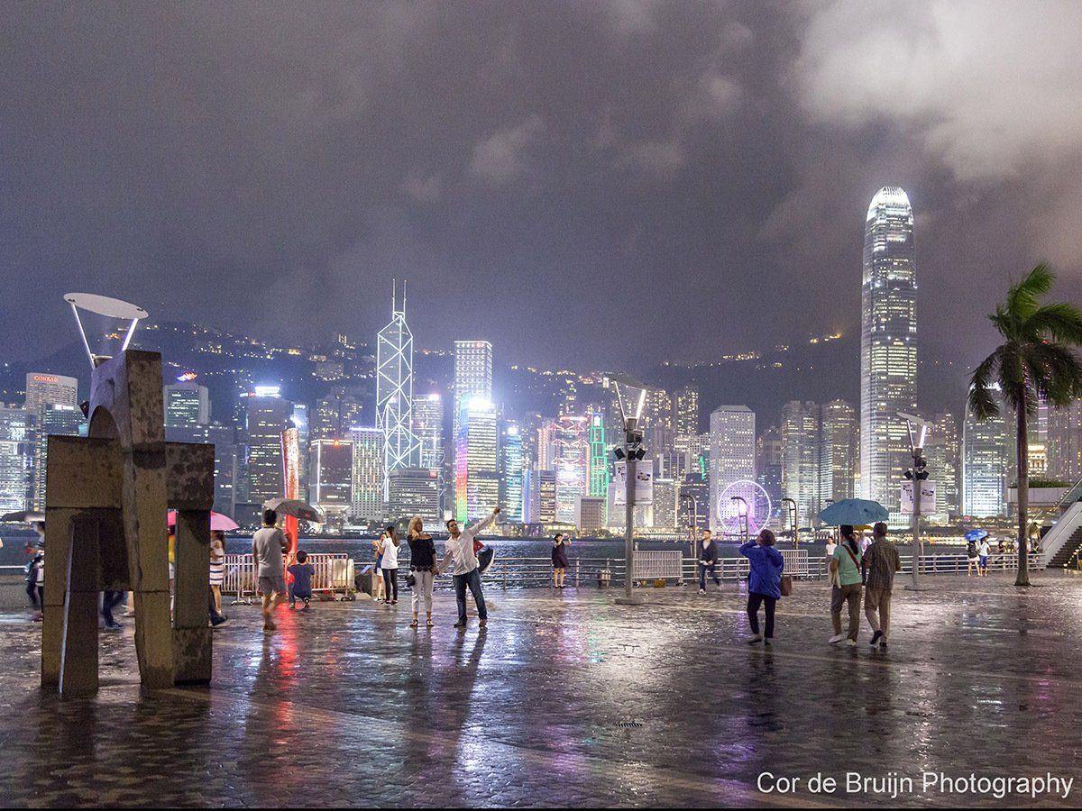 Night view of Hong Kong skyline with illuminated skyscrapers, people with umbrellas on wet pavement under dark, cloudy sky.