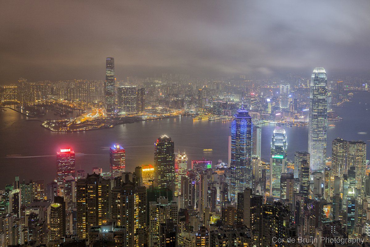 Hong Kong skyline at night, numerous illuminated skyscrapers, harbor, cloudy sky.
