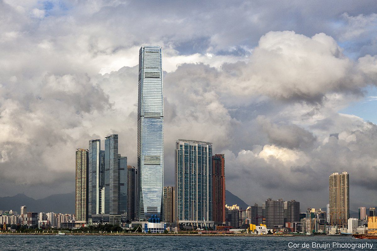 Skyscrapers in Hong Kong with a tall, modern tower in the center against a cloudy sky.