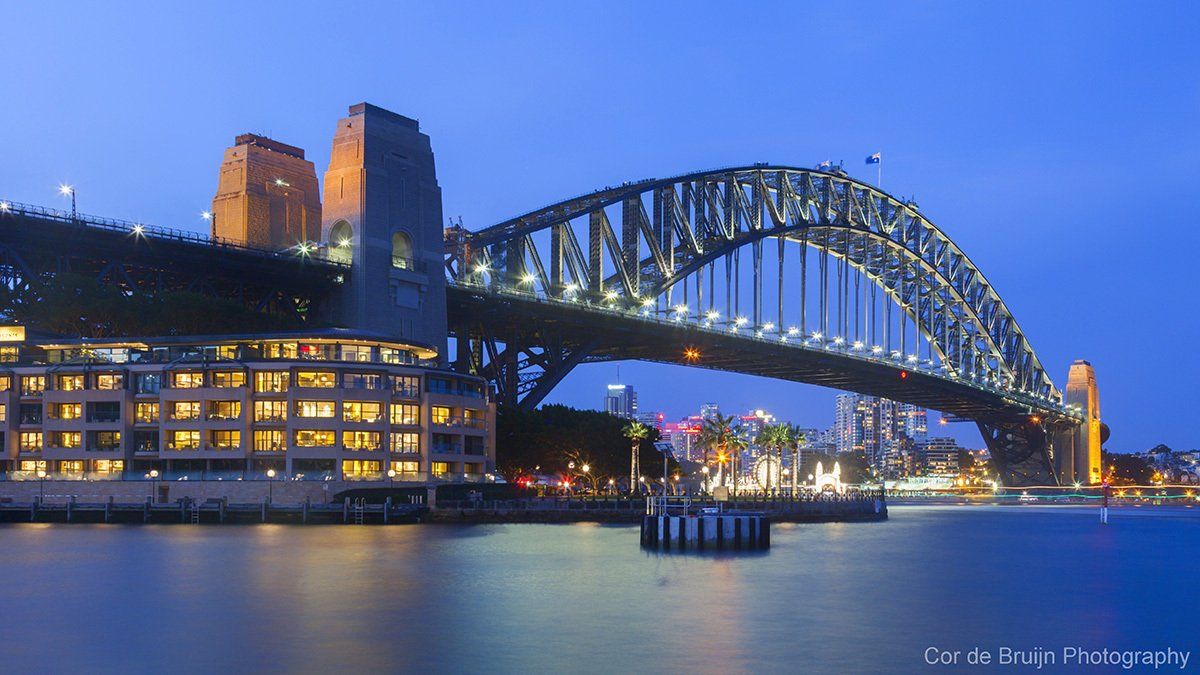 Sydney Harbour Bridge lit up at dusk over the harbor, with city buildings and reflections on the water.
