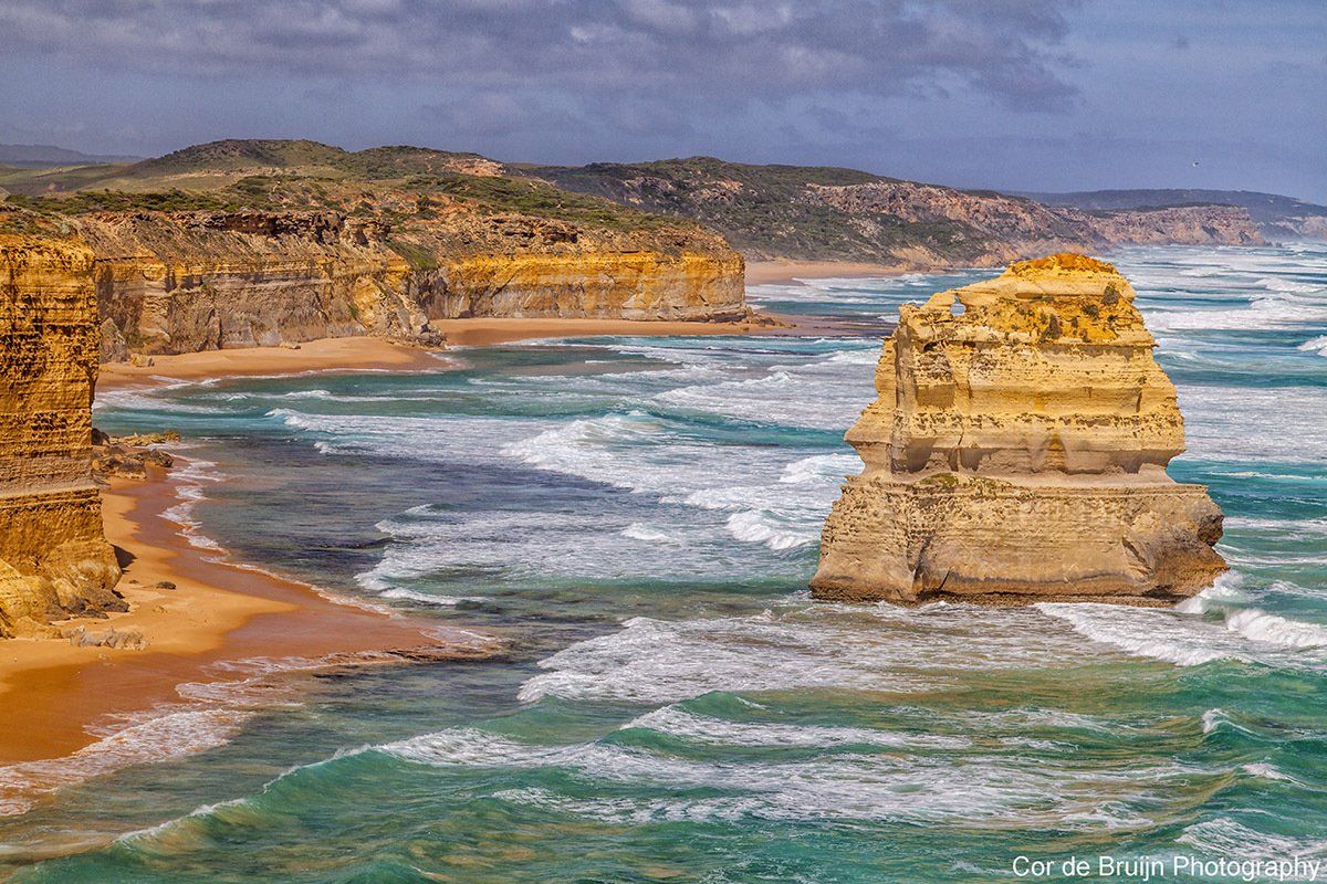Ocean waves crash against sandstone cliffs and a sea stack along a rugged coastline.