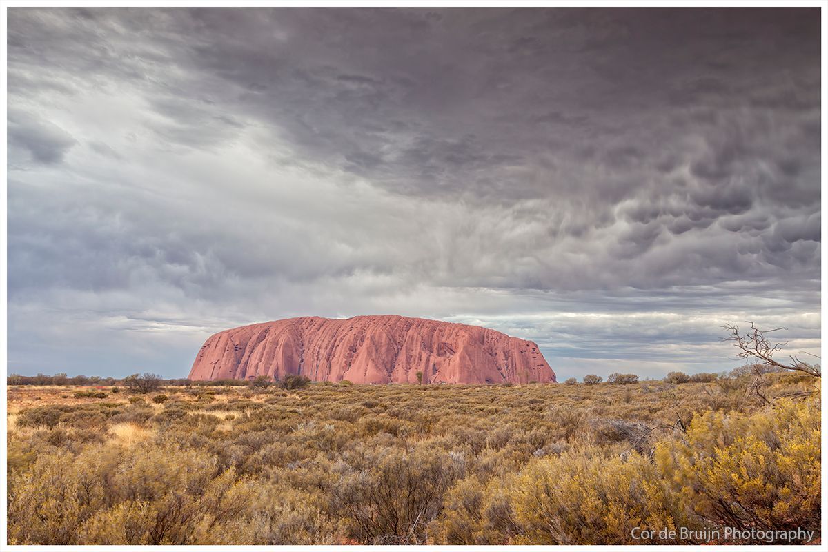Uluru monolith under a stormy sky in a desert landscape. Red rock formation, dark grey clouds.