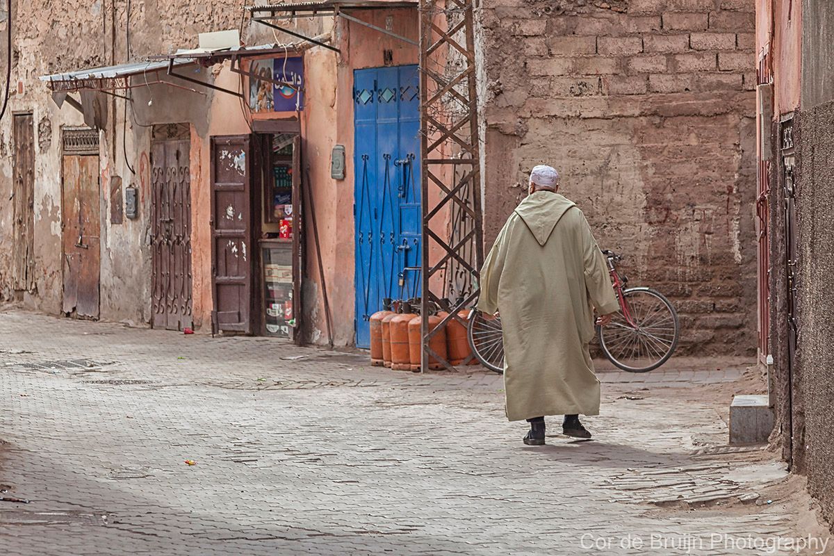 Man in traditional robe walks down a narrow alley in front of shops with blue and brown doors, Marrakech, Morocco.