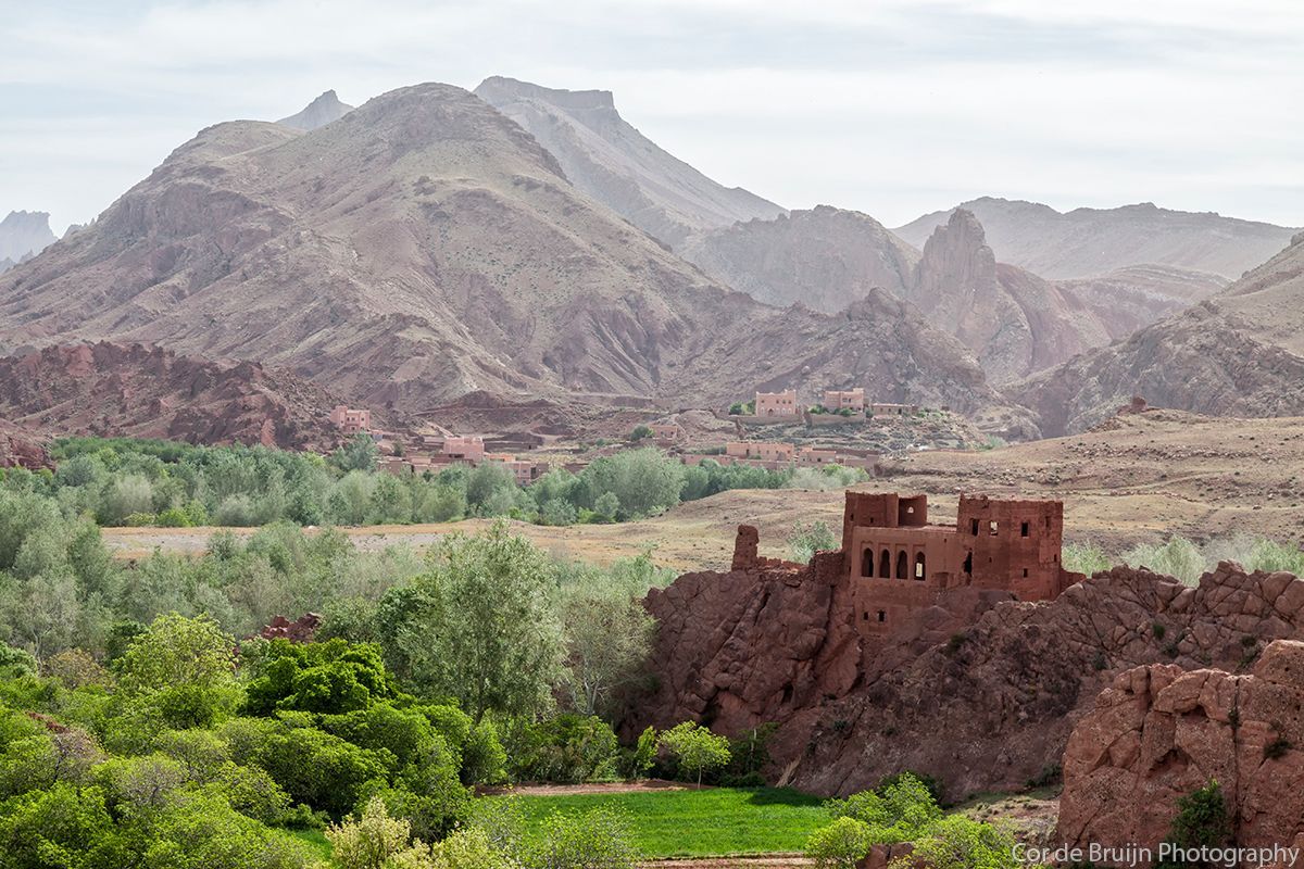 Red-hued mountains and fortress overlook a valley with green vegetation. Overcast, hazy sky.