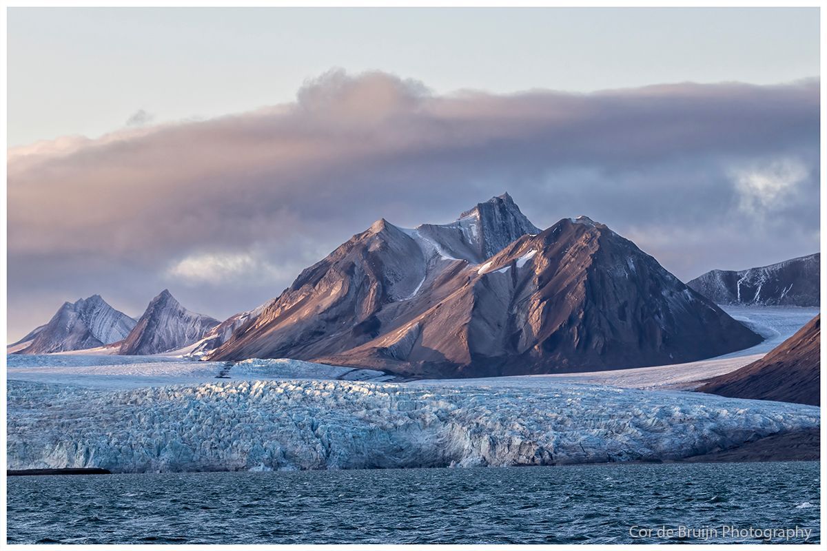 Glacier and mountains under a cloudy sky. Light blue glacier meets dark blue water.