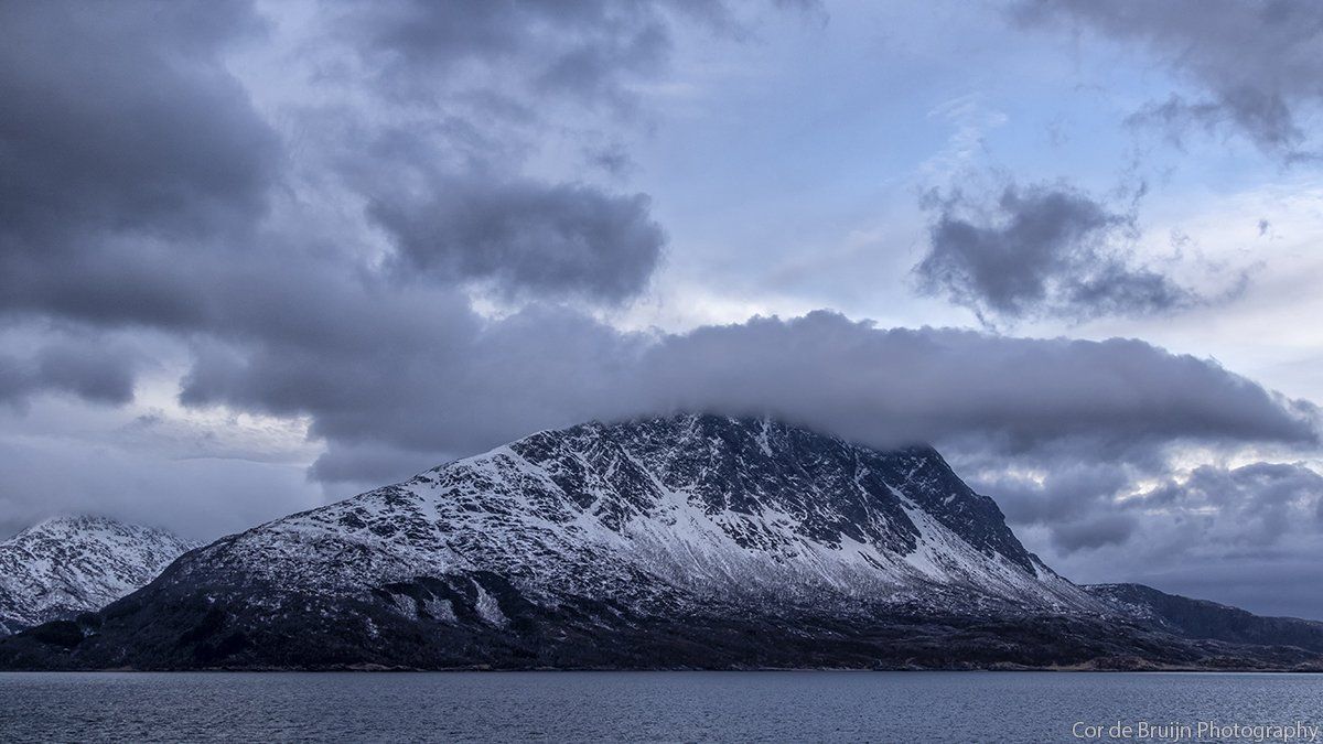 Snowy mountain peak with low clouds above water, under overcast sky.