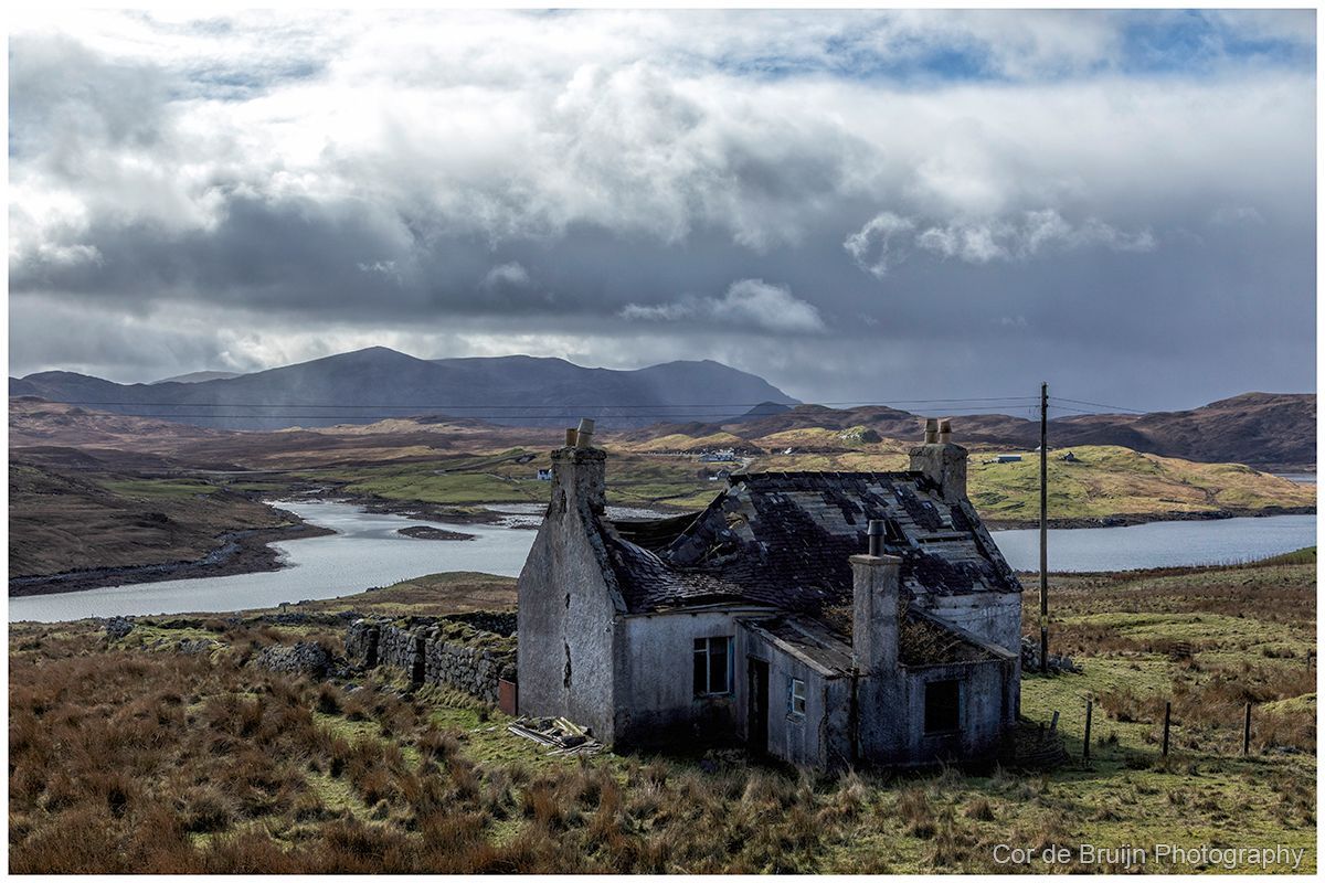 Dilapidated stone house on a hill, overlooking a lake and distant mountains under a cloudy sky.