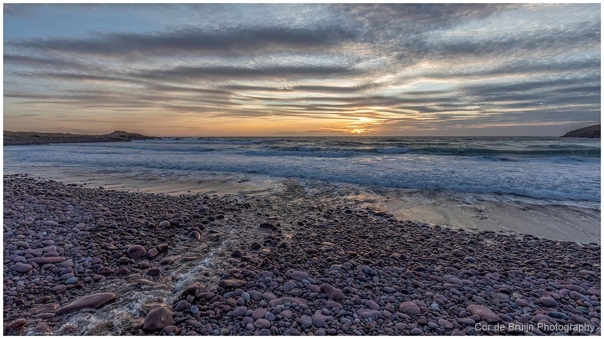 Sunset over a rocky beach with waves under a cloudy sky
