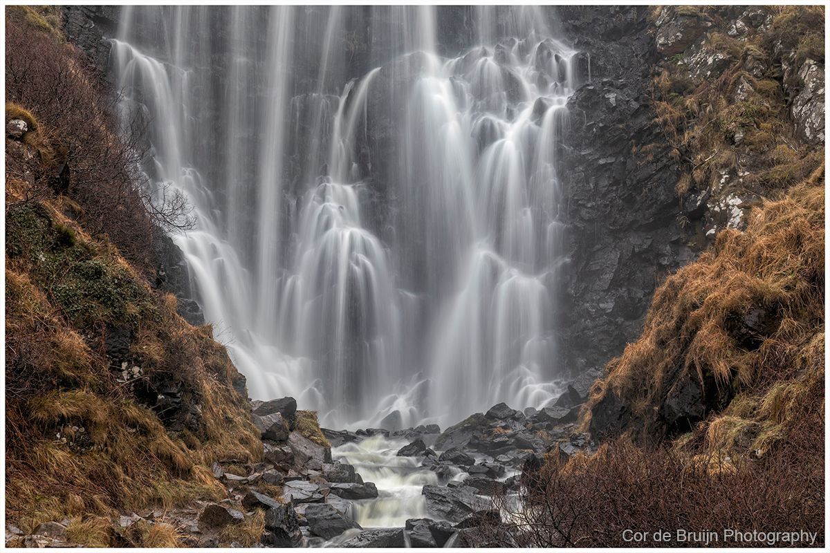 White waterfall cascading over dark rocky cliffs into a small stream in a rugged canyon.