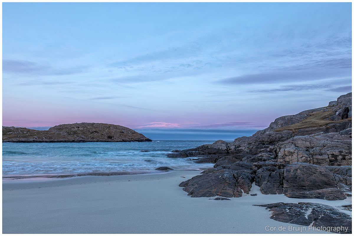 Rocky beach at dusk with calm waves, pale sand, and a pink-blue sky
